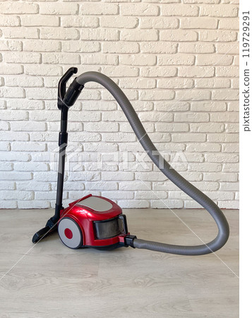 Red vacuum cleaner with a sleek design sits on a wooden floor against a white brick wall, showcasing modern home cleaning technology. Vibrant appliance contrasts with the minimalist setting 119729291