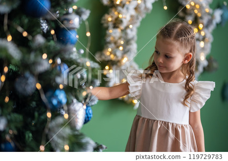 little girl in dress decorating a Christmas tree with blue and silver toys and garlands. Christmas and New Year at home 119729783
