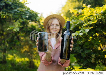 Caucasian woman vintner Shows smartphone and bottle of red wine to camera enjoying wine in her vineyard. Female in hat and an apron holding red wine bottle outdoors. Winery at autumn Caucasian woman vintner Shows smartphone and bottle of red wine to camera enjoying wine in her vineyard. Female in hat and an apron holding red wine bottle outdoors. Winery at autumn 119730201