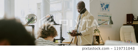 Man standing beside his desk while checking his phone in brightly lit office space with large windows and organized workstations in background Man standing beside his desk while checking his phone in brightly lit office space with large windows and organized workstations in background 119731848
