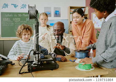 Diverse group of students gathered around a teacher explaining 3D modeling using a 3D printer in classroom setting with educational posters on walls 119731864