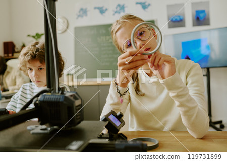 Two students engaging in hands-on learning with 3D printing technology in classroom environment. Girl closely examining a 3D-printed object while boy observes machine operation 119731899