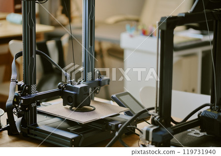 Close-up of two advanced 3D printers in a modern workspace, with an item being printed. Equipment and tools can be seen around the machines, highlighting productivity 119731940