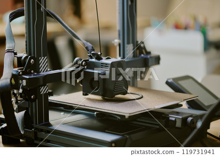 Close-up of a 3D printer during operation in a workshop setting creating an object with detailed components visible and equipment in the background. 119731941