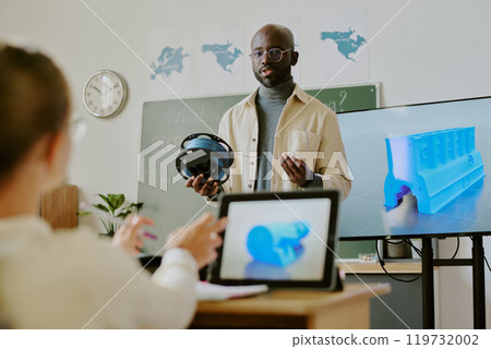 Man presenting virtual reality headset and discussing 3D model displayed on screen in classroom setting. Engaging in interactive learning environment with students observing attentively Man presenting virtual reality headset and discussing 3D model displayed on screen in classroom setting. Engaging in interactive learning environment with students observing attentively 119732002