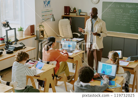 Teacher instructing students about 3D modeling on computers in classroom with various tech devices visible. Animated discussion and engagement among diverse students Teacher instructing students about 3D modeling on computers in classroom with various tech devices visible. Animated discussion and engagement among diverse students 119732025