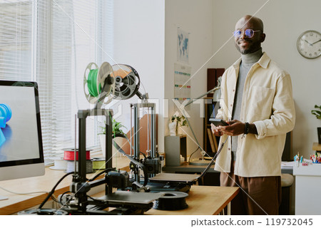 Portrait of smiling man in modern office adjusting 3D printing devices on table while standing next to window and surrounded by technology and gadgets 119732045