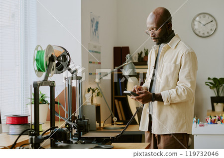 Man reading smartphone while standing in office with 3D printer and various gadgets around desk. Books and plants decorating the background 119732046