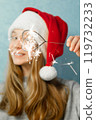 Heart shaped bengal fire in hand of beautiful girl with long red hair in New Year hat on grey background of house.  119732233