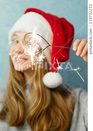 Heart shaped bengal fire in hand of beautiful girl with long red hair in New Year hat on grey background of house. Heart shaped bengal fire in hand of beautiful girl with long red hair in New Year hat on grey background of house. 119732233