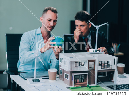 Engineers meeting about wind turbine model and house model. Renewable energy and housing development concept. Team of architects and engineers in modern office discussing design structure. 119733225
