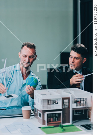 Engineers meeting about wind turbine model and house model. Renewable energy and housing development concept. Team of architects and engineers in modern office discussing design structure. Engineers meeting about wind turbine model and house model. Renewable energy and housing development concept. Team of architects and engineers in modern office discussing design structure. 119733226
