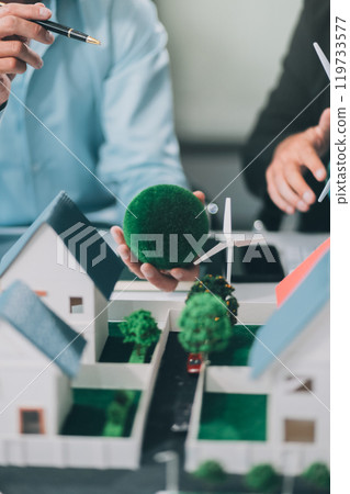 Engineers meeting about wind turbine model and house model. Renewable energy and housing development concept. Team of architects and engineers in modern office discussing design structure. 119733577