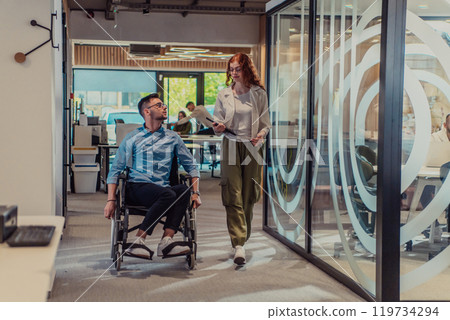Young business colleagues, collaborative business colleagues, including a person in a wheelchair, walk past a modern glass office corridor, illustrating diversity, teamwork and empowerment in the Young business colleagues, collaborative business colleagues, including a person in a wheelchair, walk past a modern glass office corridor, illustrating diversity, teamwork and empowerment in the 119734294