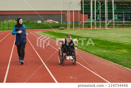 A Muslim woman in a burqa running together with a woman in a wheelchair on the marathon course, preparing for future competitions. 119734448