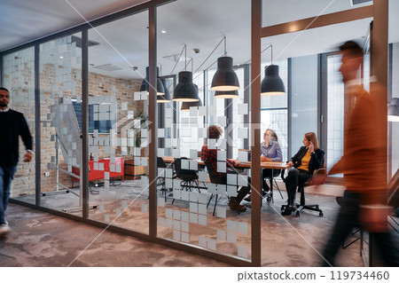 In the dynamic hustle and bustle of a business environment, a group of young business professionals walk down the corridor next to their office, while their colleagues collaborate inside, symbolizing 119734460
