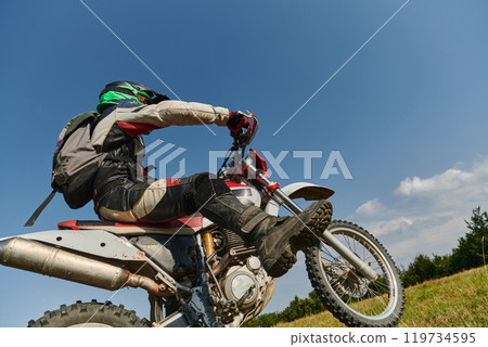 A fearless professional motocross rider skillfully executes an extreme back wheelie maneuver through a perilous forest terrain, showcasing remarkable balance and daring expertise 119734595