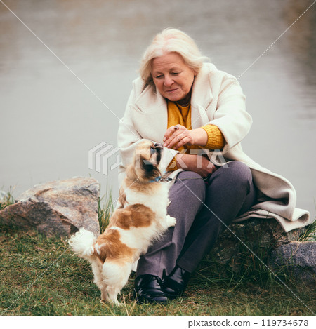 Cozy autumn picnic. Elderly woman sitting on blanket by the river, feeding her dog, spending lovely time with her dog. 119734678
