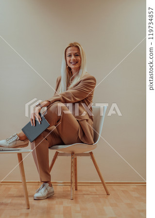 A professional businesswoman sits on a chair, surrounded by a serene beige background, diligently working on her laptop, showcasing dedication and focus in her pursuit of success A professional businesswoman sits on a chair, surrounded by a serene beige background, diligently working on her laptop, showcasing dedication and focus in her pursuit of success 119734857