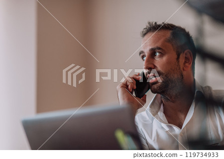 A businessman talking on his smartphone while seated in an office, showcasing his professional demeanor and active communication. A businessman talking on his smartphone while seated in an office, showcasing his professional demeanor and active communication. 119734933
