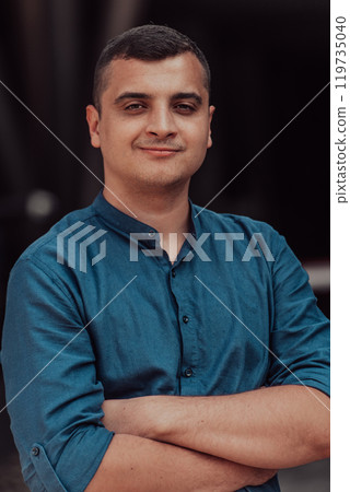 A successful young businessman in a shirt, with crossed arms, poses outdoors, confident expression on his face. 119735040