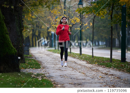 Young beautiful woman running in autumn park and listening to music with headphones on smartphone Young beautiful woman running in autumn park and listening to music with headphones on smartphone 119735096