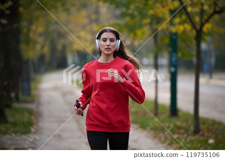Young beautiful woman running in autumn park and listening to music with headphones on smartphone 119735106