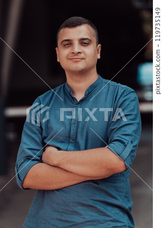 A successful young businessman in a shirt, with crossed arms, poses outdoors, confident expression on his face. A successful young businessman in a shirt, with crossed arms, poses outdoors, confident expression on his face. 119735149