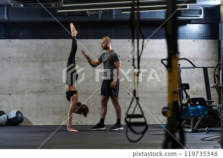 A muscular man assisting a fit woman in a modern gym as they engage in various body exercises and muscle stretches, showcasing their dedication to fitness and benefiting from teamwork and support 119735348