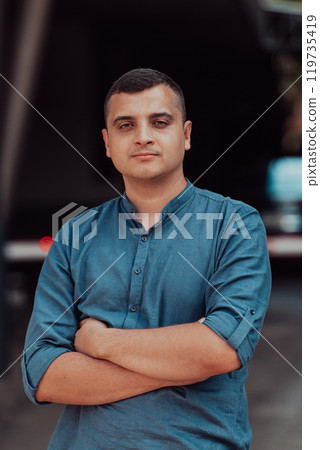 A successful young businessman in a shirt, with crossed arms, poses outdoors, confident expression on his face. 119735419