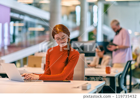 In a modern startup office, a professional businesswoman with orange hair sitting at her laptop, epitomizing innovation and productivity in her contemporary workspace. 119735432
