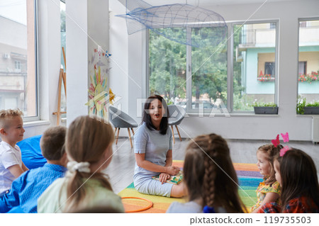 A happy female teacher sitting and playing hand games with a group of little schoolchildren 119735503