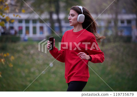 Young beautiful woman running in autumn park and listening to music with headphones on smartphone Young beautiful woman running in autumn park and listening to music with headphones on smartphone 119735679