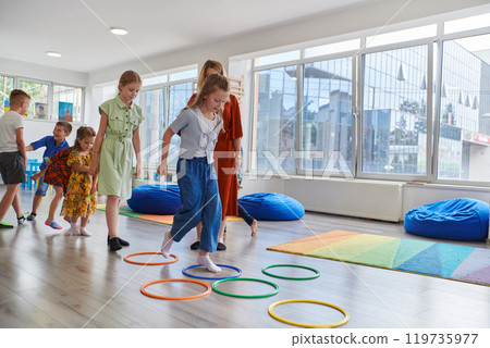 Small nursery school children with female teacher on floor indoors in classroom, doing exercise. Jumping over hula hoop circles track on the floor. Small nursery school children with female teacher on floor indoors in classroom, doing exercise. Jumping over hula hoop circles track on the floor. 119735977