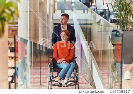 A company director assists his business colleague in a wheelchair, helping her navigate to their startup office, where they work alongside their diverse team of colleagues, emphasizing inclusivity and A company director assists his business colleague in a wheelchair, helping her navigate to their startup office, where they work alongside their diverse team of colleagues, emphasizing inclusivity and 119736317