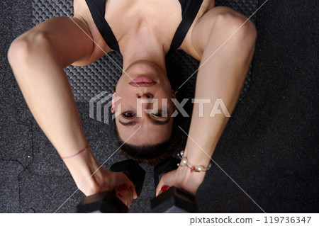 A fit woman is lying on the gym floor, performing arm exercises with dumbbells and showcasing her dedication and strength. 119736347
