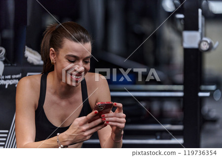 A fit woman in the gym taking a break from her training and uses her smartphone, embracing the convenience of technology to stay connected 119736354