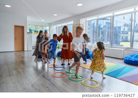 Small nursery school children with female teacher on floor indoors in classroom, doing exercise. Jumping over hula hoop circles track on the floor. 119736427