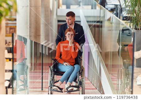 A company director assists his business colleague in a wheelchair, helping her navigate to their startup office, where they work alongside their diverse team of colleagues, emphasizing inclusivity and 119736438