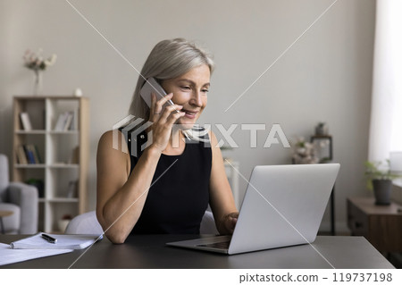 Busy older woman speak on cell check data on laptop Busy older woman speak on cell check data on laptop 119737198
