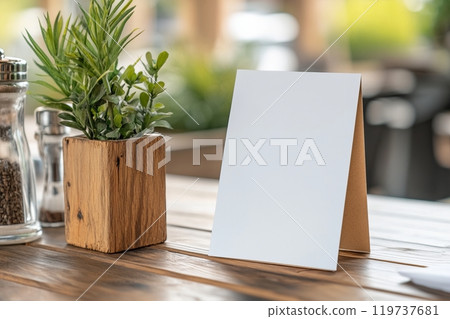 Blank table tent card placed on a wooden table beside a potted plant in a cozy outdoor cafe 119737681