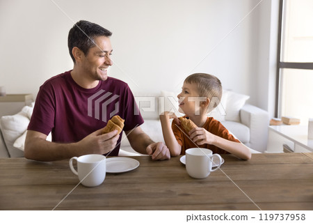 Happy Hispanic dad and positive cute son child eating croissants Happy Hispanic dad and positive cute son child eating croissants 119737958