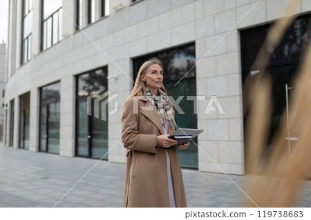 A businesswoman in a tan coat stands outside a business center, holding a tablet and looking 119738683