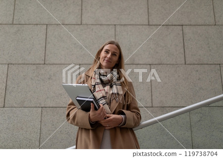 A businesswoman stands near a business center, holding a laptop and books, looking confident A businesswoman stands near a business center, holding a laptop and books, looking confident 119738704