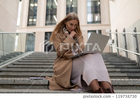 A business woman works on her laptop while seated on the steps of a modern office building during a 119738730