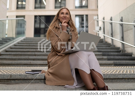 A business woman working thoughtfully on her laptop while seated on the steps of a modern business A business woman working thoughtfully on her laptop while seated on the steps of a modern business 119738731