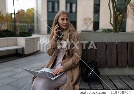 A businesswoman sits on a bench outside a business center, talking on her phone and working on her 119738764