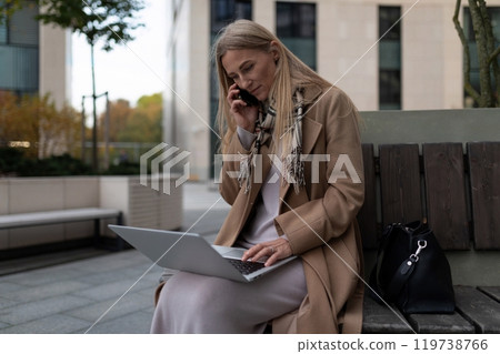 A businesswoman sits on a bench outside a business center, working on her laptop and talking on her A businesswoman sits on a bench outside a business center, working on her laptop and talking on her 119738766