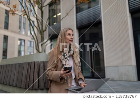 A businesswoman sits on a bench outside a business center, working on her laptop and checking her 119738767
