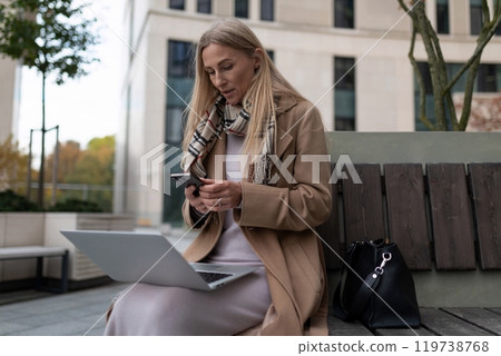 A businesswoman uses her phone while sitting on a bench outside a business center 119738768